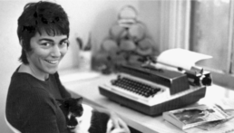 Black and white image of a smiling woman with short dark hair seated in front of a typewriter