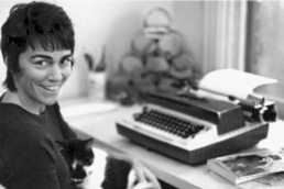 Black and white image of a smiling woman with short dark hair seated in front of a typewriter